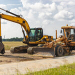The excavator and the grader stand side by side against the blue sky. Heavy construction earthmoving equipment. Construction of roads and underground communications. Construction industry.